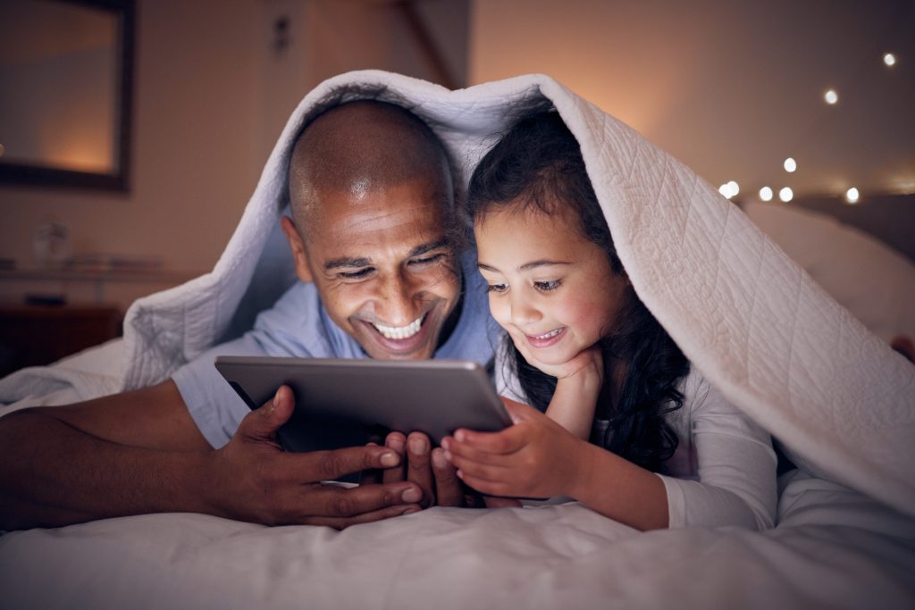 Father, kid and smile with tablet at night under blanket of online games, reading ebook or storytelling app\. Happy dad, girl child or watch cartoon movie on digital tech, media or internet in bedroom\.