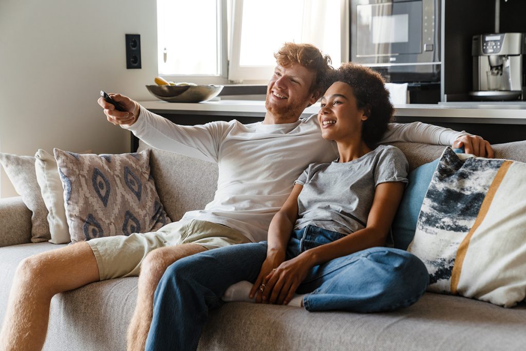 Young beautiful happy smiling interracial couple watching tv , while sitting on couch at home together\. Handsome redhead man and beautiful smiling african woman