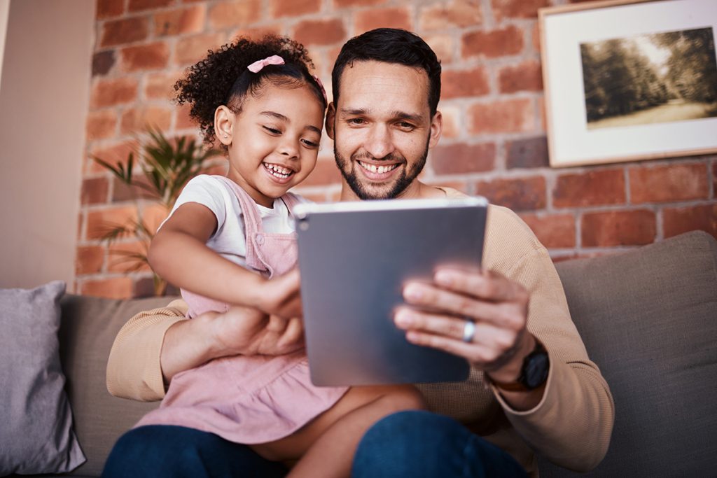 Dad, child and tablet for learning on sofa in family home or reading online, playing a game or technology for education\. Father, kid and mobile app to teach or streaming cartoon and relax watching tv\.