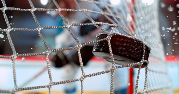 Close\-up action shot of an ice hockey puck hitting the goal net during a scoring moment\. Dynamic sports scene with ice spray and a blurred hockey player in the background capturing the intensity of the game\.